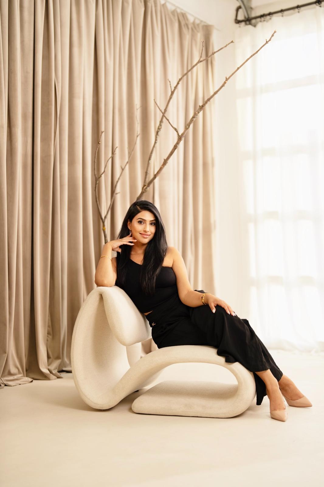 Kiran Gandhi seated on a cream sculptural chair, beige drapery and bare branch arrangement